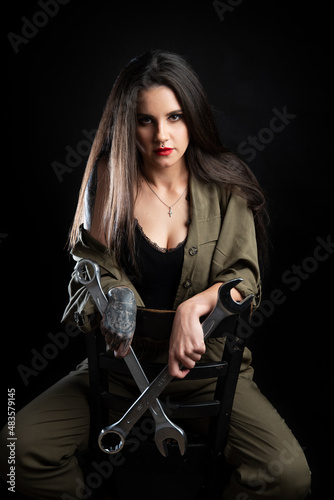 Piękna dziewczyna z tatuażami przebrana w strój mechanika, siedzi na krześle i trzyma bardzo duże klucze. Zdjęcie zrobione w studio na czarnym tle, oświetlenie z 2 lamp.