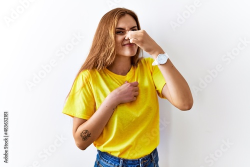 Young brunette woman standing over isolated background smelling something stinky and disgusting, intolerable smell, holding breath with fingers on nose. bad smell