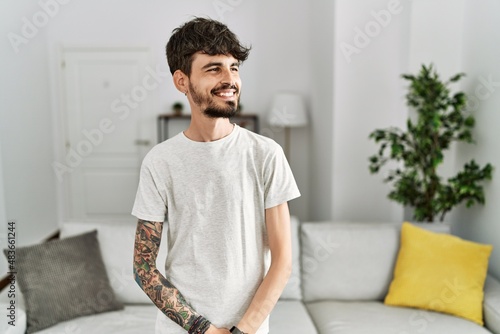 Hispanic man with beard at the living room at home looking away to side with smile on face, natural expression. laughing confident.