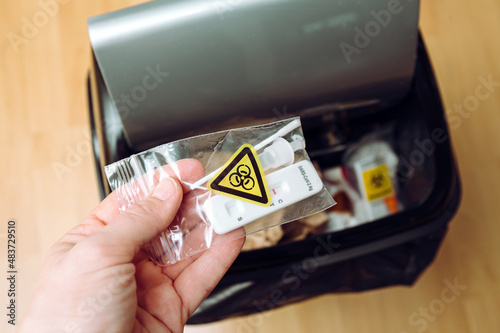 Safe handling and management of rapid antigen COVID-19 testing waste concept. Person hand throw away pieces of antigen test kit sealed in plastic bag, biohazard symbol. Home use.
