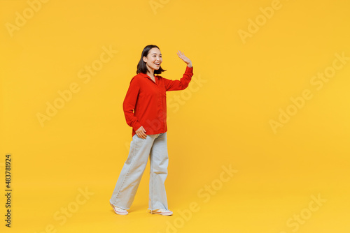 Full size body length cheerful young woman of Asian ethnicity 20s in casual clothes look camera go move meet greet waving hand as notices someone isolated on plain yellow background studio portrait.
