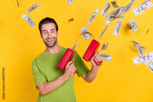 Photo of joyful beard millennial guy with money gun wear green t-shirt isolated on yellow color background