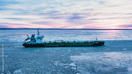 Enormous cargo ship sailing through ocean water with thick layer of broken ice pieces covering surface on polar day aerial view