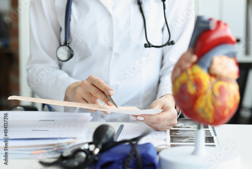 Doctor cardiologist examining cardiogram against background of artificial heart model closeup