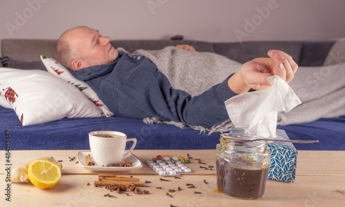 Mężczyzna z gorączką leży w łóżku. Na stole tabletki, herbata, chusteczki na katar, cytryna i miód.