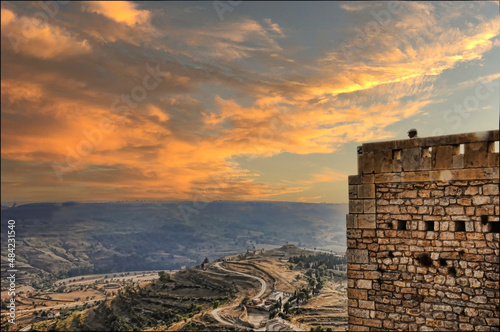 Vista desde lo alto de un valle en España con un camino descendente. Un atardecer de nubes rojizas y al costado una alta pared de piedras con la cabeza sobresaliente de una persona no identificable.