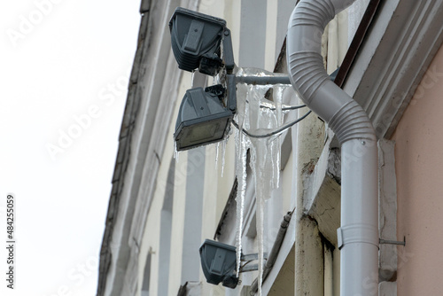 A lighting device is installed on the wall of the building. Icicles and snow hang on the spotlight. Winter utopian urban landscape with a view of frozen buildings.