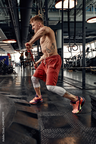 Strength concept. Male athlete using hammer during fitness workout. Fitness man beating rubber tire in gym. Muscular sportsman performing intensity training with sledgehammer in sport club