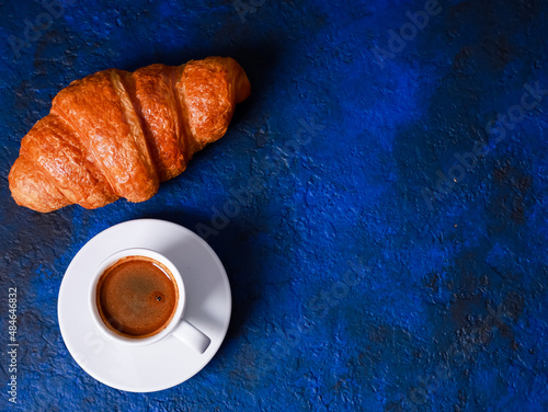 Fresh baked croissant, white mug of expresso on blue concrete table. Cup of hot coffee, buns, rolls close up. Food, French breakfast, morning menu, cafe concept. Top view, copy space