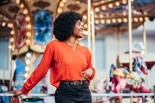 Smiling dreamy young black woman with afro hairstyle and red sweater standing next to a carousel on the street