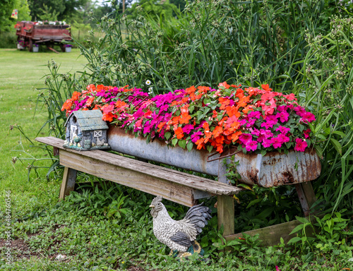 An old chicken feeder filled with colorful impatiens and decorative rural ornaments and an Amish farm wagon sits in the background. 