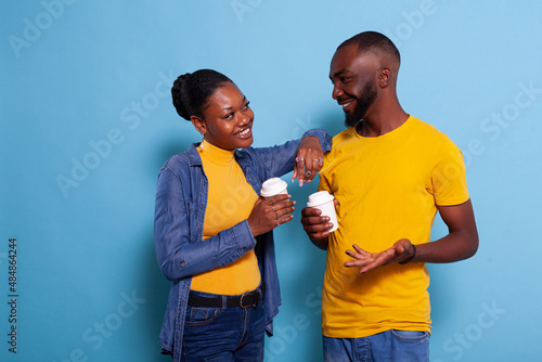 Romantic couple in love looking at each other in studio, holding cup of coffee in hand. Girlfriend and boyfriend flirting and smiling, standing over blue background and enjoying drink.