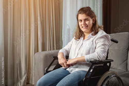 Young disabled woman with infantile cerebral paralysis in white shirt smiles sitting in wheelchair near stylish sofa in living room at home, sunlight.