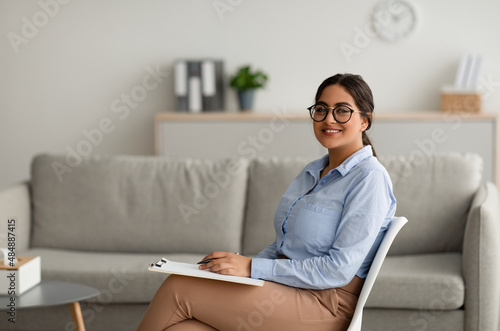 Confident arab female psychologist writing in clipboard, sitting at modern office and smiling at camera, free space