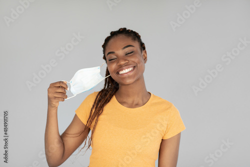 End of coronavirus pandemic. Happy black woman taking off medical mask, feeling free on grey studio background