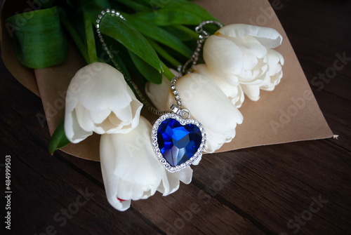 Close-up of the Heart of The Ocean necklace with white tulips bouquet on a craft paper. Titanic necklace. Blue diamond in a heart-shaped. Diamond necklace.