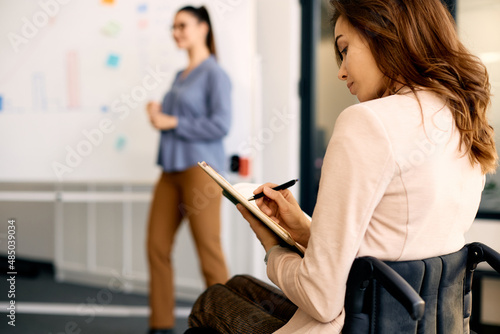 Young businesswoman in wheelchair takes notes while attending seminar in the office.