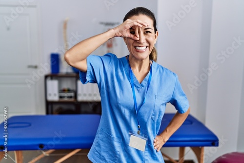 Young hispanic woman wearing physiotherapist uniform standing at clinic doing ok gesture with hand smiling, eye looking through fingers with happy face.