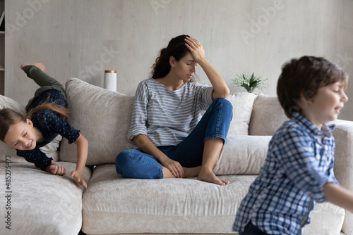 Tired young mother suffering from headache, feeling annoyed and exhausted by noisy kids running around, upset mom or babysitter sitting on couch, having problem with naughty active children