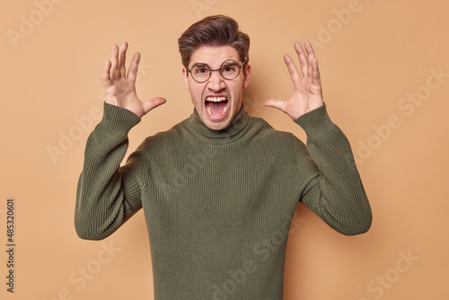 Photo of outraged man yells from annoyance screams loudly keeps palms raised expresses negative emotions wears round spectacles and sweater isolated over beige background. Furious male model