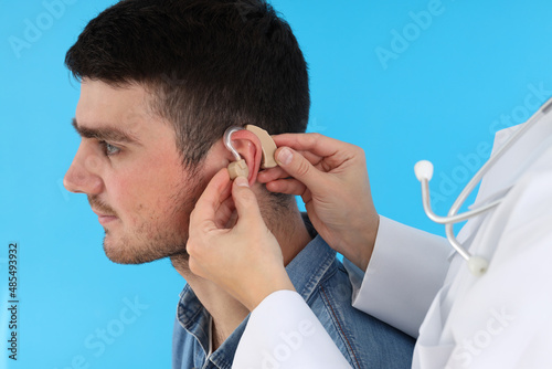 Doctor installs hearing aid on young man ear on blue background