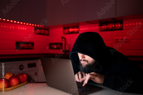 a hacker in a black hoodie with a hood on his head is sitting at a laptop trying to hack a social network