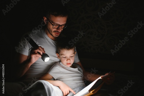 Father and son with flashlight reading book under blanket at home