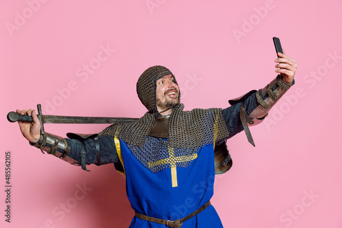Half-length portrait of man, medieval warrior or knight in protective chain armor with sword taking selfie on phone isolated over pink background