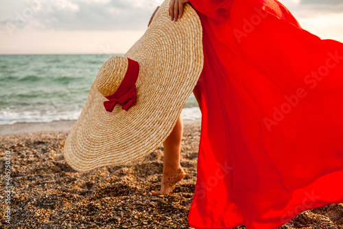 Beautiful strong woman wearing maxi straw hat and red chiffon cape like dresss walking in the beach against the blue sky and sea