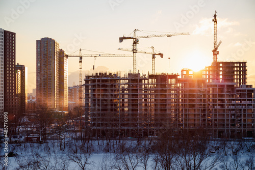Lots of tower cranes build high-rise residential neighborhood on bank of frozen river in winter. Industrial concept with sun flare. Yellow light and blue shadows.