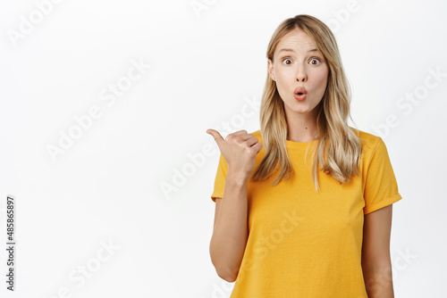 Surprised young woman pointing left with curious expression, interesting thing near, standing in t-shirt over white background