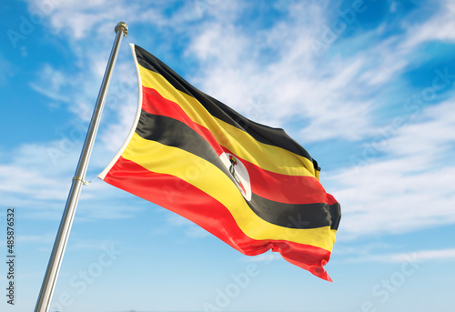 3d rendering Uganda flag waving in the wind on flagpole. Perspective wiev Uganda flag waving a blue cloudy sky