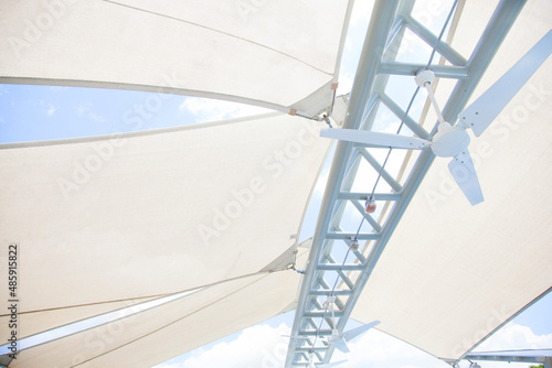 Looking up at a bright white outdoor canvas canopy and geometric architectural strut with a ceiling fan, protecting patio from the summer sun and bright blue sky
