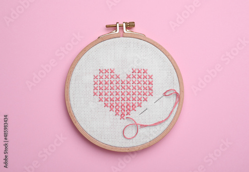 Canvas with embroidered heart and needle in hoop on pink background, top view