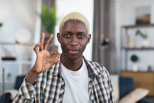 Portrait of african man with blond hair and blue eyes gesturing sign OK with fingers while sitting at home and looking at camera. Concept of people and approvement.