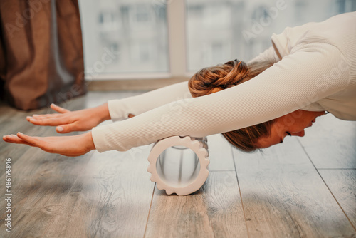 Adult athletic woman, in white bodysuit, performing fascia exercises on the floor - caucasian woman using a massage foam roller - a tool to relieve tension in the back and relieve muscle pain - the