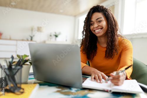 Woman sitting at desk, using computer and writing in notebook