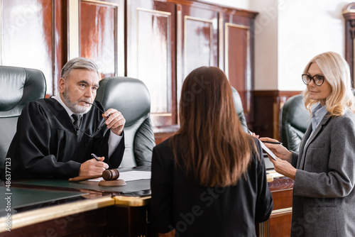 thoughtful judge holding eyeglasses listen to prosecutor standing near attorney in court