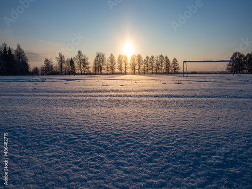 Snow sparkles at snowy football field in winter evening. Silhouette of trees in rays of the setting sun. Place for your text. The cold season.