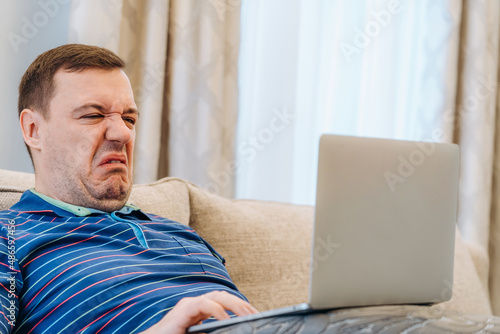 Annoyed millennial man with disgust confused looking at laptop screen sitting on sofa at living room. cringe guy looking at computer with squeamishness, reacting on watching disgusting video at home