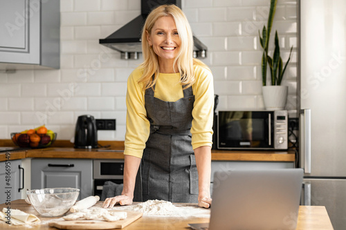 Charming good-looking middle-aged housewife in apron cooking at home, serene mature woman in apron standing in modern kitchen, looking at the camera and siles