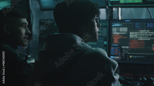Young male hacker in glasses looking at screen and talking with associate while performing cyber attack on computer in dim room of criminal base