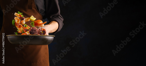 A professional chef in a dark uniform prepares assorted seafood - pieces of red fish, octopus, shrimp in a pan on a black background. Levitation. Sea oriental, Thai cuisine.
