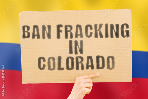 The phrase " Ban fracking in Colorado " on a banner in men's hand with blurred Colorado flag on the background. Nature. Extract. Stop. Pipe. Energy. Drill. Raw. Power. Fuel. Fossil. Crisis