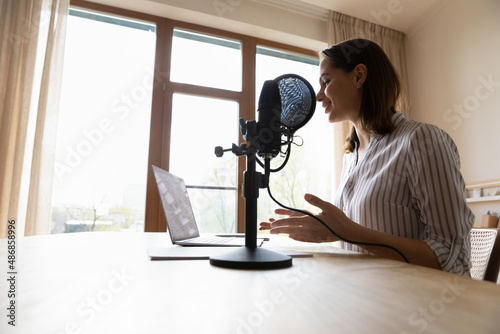 Smiling professional female radio host talking in stand microphone, looking at laptop screen, voice acting online, streaming live video lecture or educational webinar, vlogging at home office.