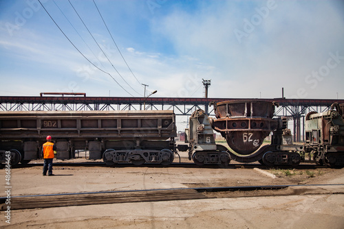 Metallurgical plant (smelter) station. Train with rusted slag car wagon. Switcher worker in orange safety jacket on foreground. Blue sky background.
