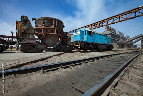 Metal alloys plant. Blue locomotive and rusted slag cars. Metallurgical plant (smelter) main industrial building on background. Blue sky.
