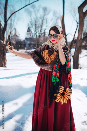 Outdoors lifestyle portrait of pretty woman in a traditional Russian folk clothes with bagels on winter background. Taking selfie. Wearing stylish retro sunglasses. Maslenitsa. Stylization old times