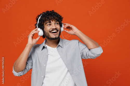 Cheerful exultant happy young bearded Indian man 20s years old wears blue shirt listen music in headphones dance have fun gesticulating hands relax isolated on plain orange background studio portrait.