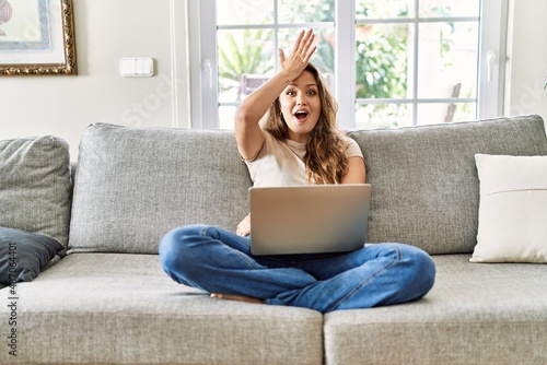 Beautiful young brunette woman sitting on the sofa using computer laptop at home surprised with hand on head for mistake, remember error. forgot, bad memory concept.